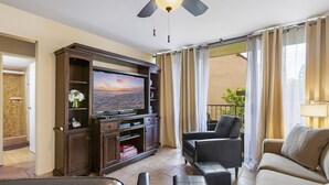 A hotel living room with a flat-screen TV, leather seating, a wooden entertainment unit, and a balcony with curtains.