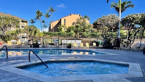 A swimming pool with a tiled deck and lounge chairs, surrounded by palm trees and a building in the background.