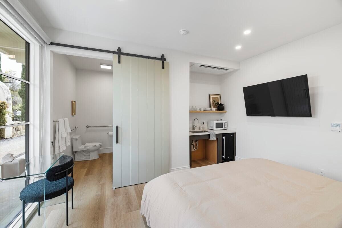 A modern bedroom with a bed, a TV mounted on the wall, a bathroom with a toilet and sink, and a sliding door leading to a balcony.