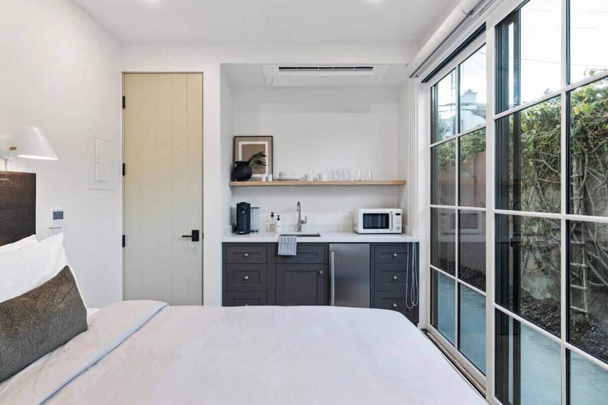 A modern bedroom with a bed, a bedside lamp, a small kitchenette with a microwave, and a large window offering a view of greenery.