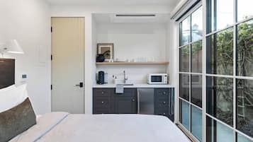 Een moderne slaapkamer met een bed, een bedlamp, een kleine kitchenette met een magnetron en een groot raam met uitzicht op groen.