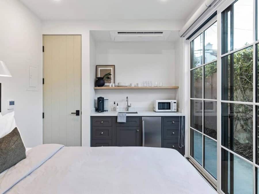 A modern bedroom with a bed, a bedside lamp, a small kitchenette with a microwave, and a large window offering a view of greenery.