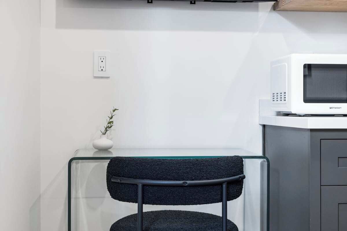 A modern kitchen with a white microwave on a white countertop, a glass table, and a dark chair with a metal frame.