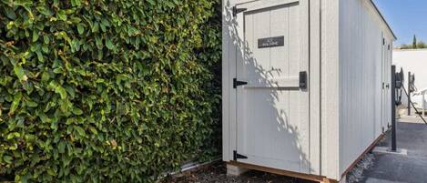 Um depósito de ferramentas branco com uma porta identificada como 'AIR MACHINE' ao lado de um cercado verde denso.