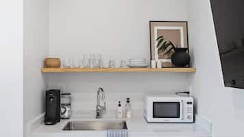 Uma cozinha compacta com bancada branca, pia de aço inoxidável, micro-ondas, cafeteira e prateleira com copos e uma moldura com uma fotografia.