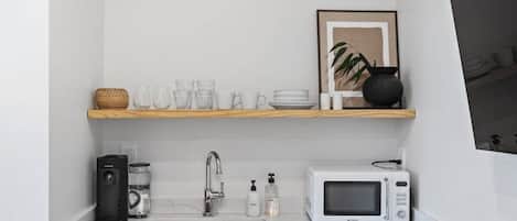 A compact kitchen with a white countertop, a stainless steel sink, a microwave, a coffee maker, and a shelf with glassware and a framed picture.
