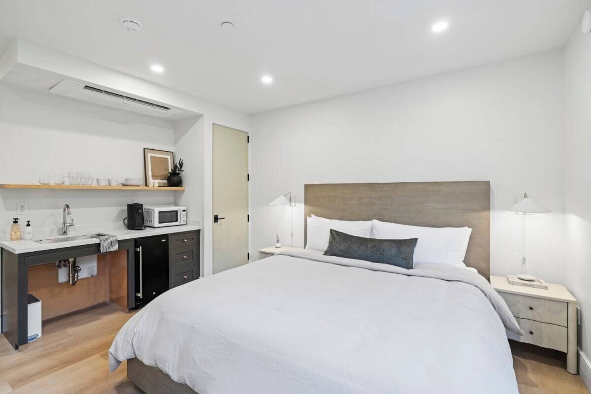 A modern hotel room with a bed, bedside tables, a desk with a microwave, and a small kitchenette.
