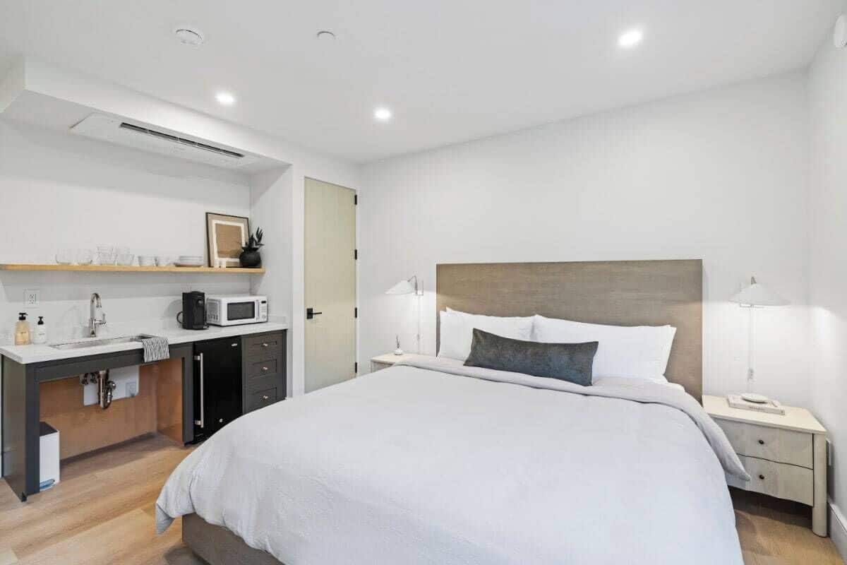 A modern hotel room with a bed, bedside tables, a desk with a microwave, and a small kitchenette.