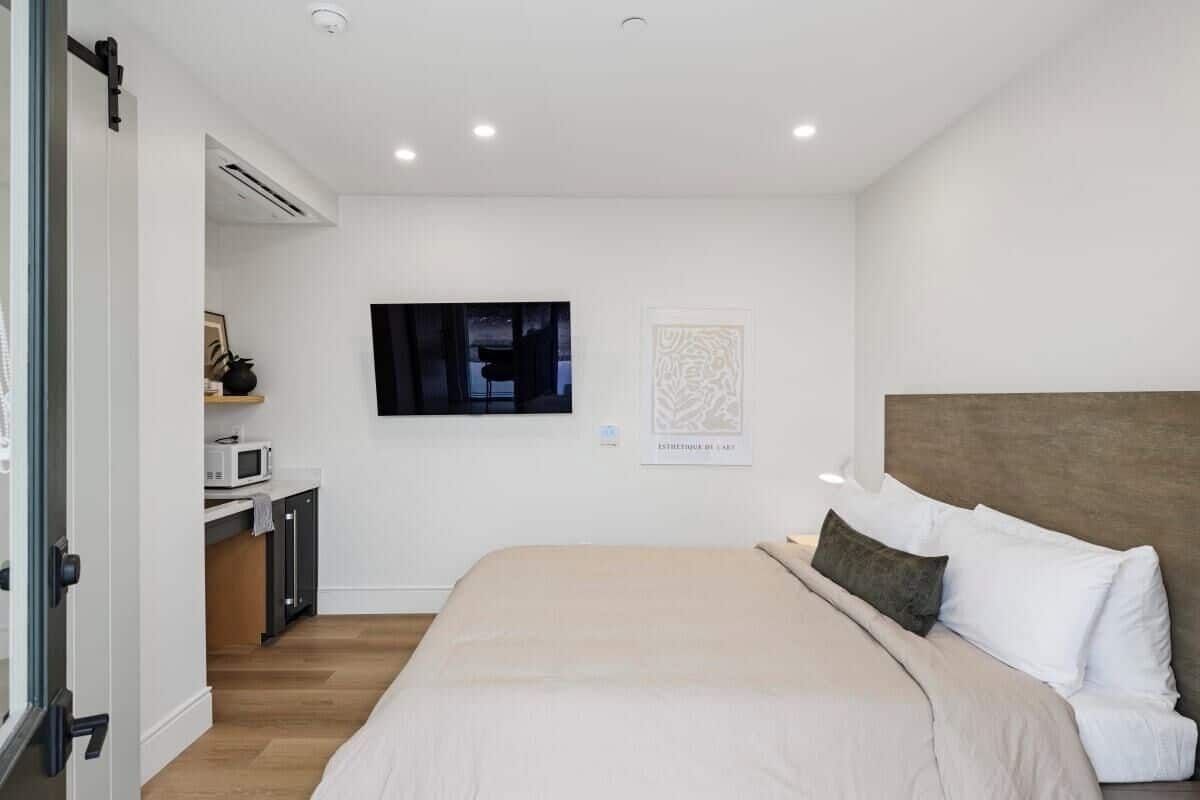 A modern bedroom with a large bed, a flat-screen TV mounted on the wall, a small kitchenette, and a door leading to another room.