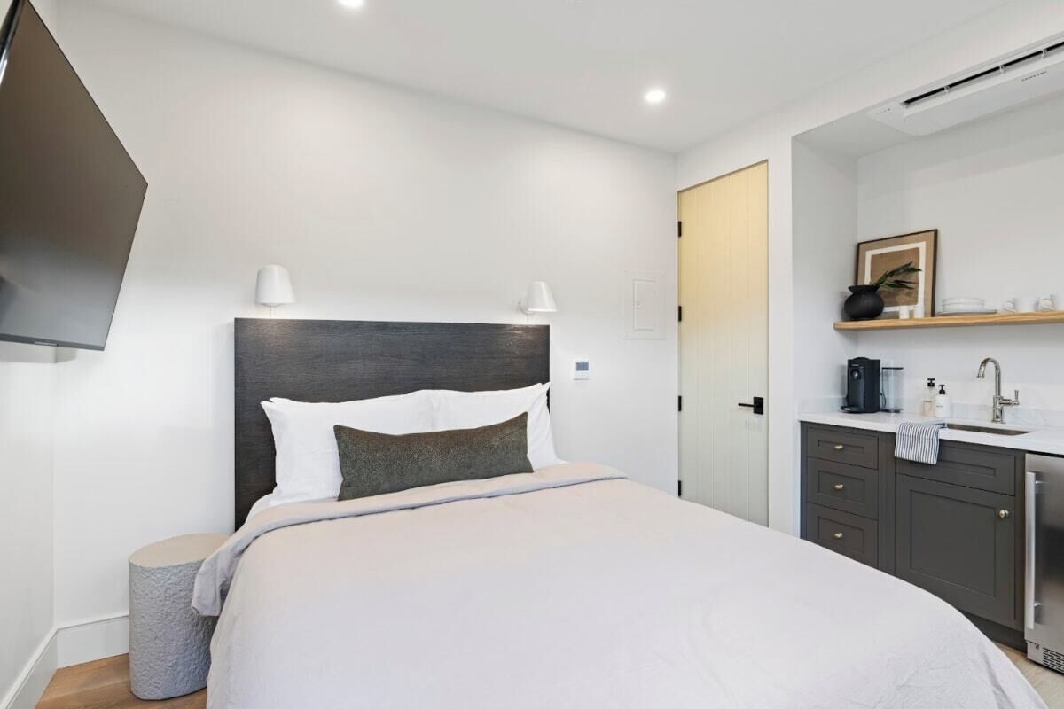 Une chambre d’hôtel moderne avec un grand lit, des lampes de chevet, une armoire intégrée, une petite table ronde et un miroir au-dessus du lavabo.