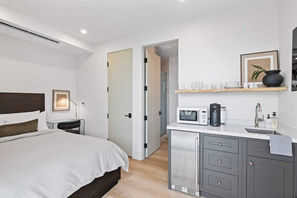 A modern hotel room with a bed, bedside table, and a kitchenette area with a microwave, coffee maker, and sink.