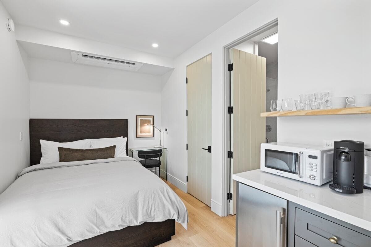 A modern bedroom with a bed, bedside table, and a kitchenette area with a microwave and coffee maker.