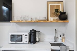 A kitchen with a microwave, coffee maker, and sink. There are shelves above holding glasses, plates, and a framed picture. - Casa Jardin, a ZenStay Hotel (Santa Barbara)