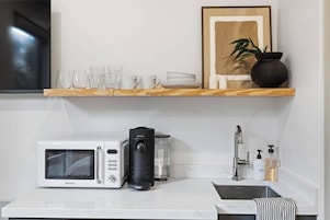 Cozinha equipada com micro-ondas, máquina de café e pia. Há prateleiras com copos, pratos e um quadro emoldurado.