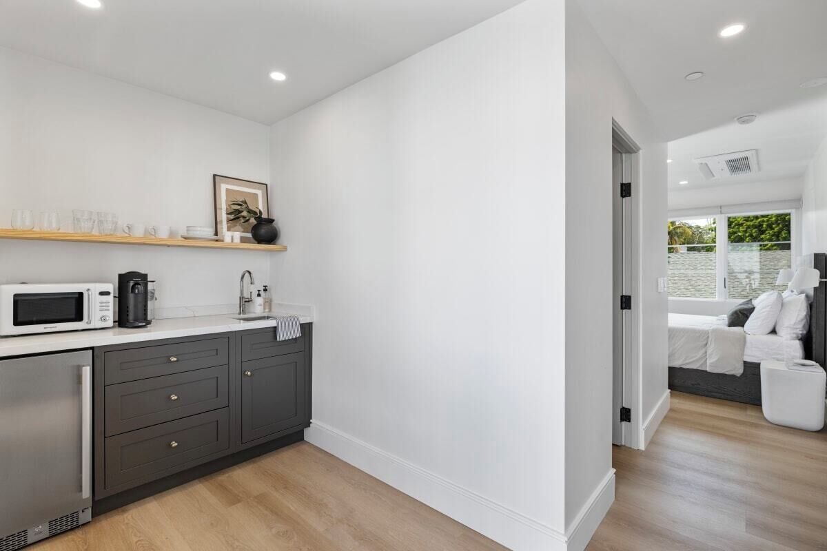 A modern kitchen with dark cabinets, a white countertop, and a microwave. There is a wooden shelf above the counter holding cups and a framed picture.