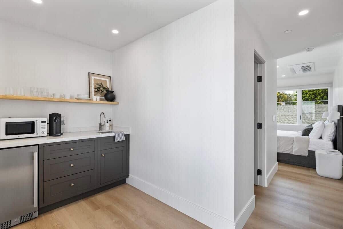 A modern kitchen with dark cabinets, a white countertop, and a microwave. There is a wooden shelf above the counter holding cups and a framed picture.