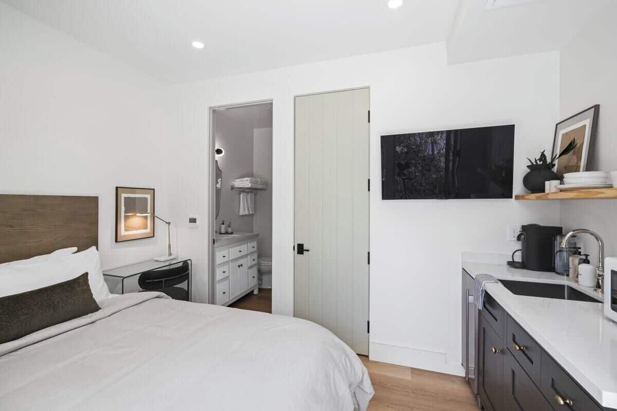 A modern hotel room with a large bed, a desk, a vanity, and a kitchen area.