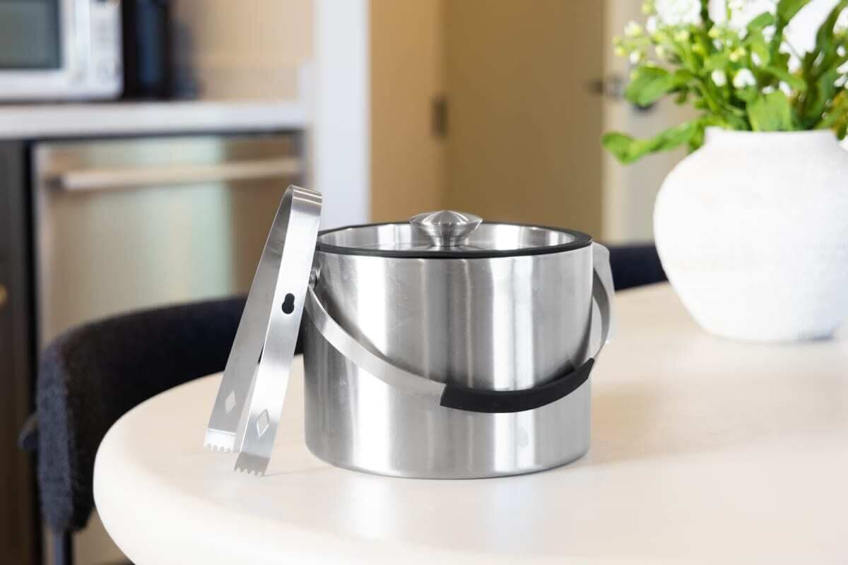 A stainless steel pot on a white table with a black handle, a white vase with green plants, and a microwave in the background.