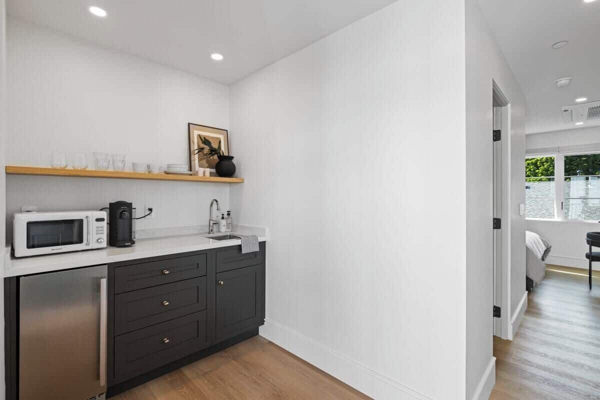 A modern kitchen with a microwave, coffee maker, and sink. There is a wooden shelf above the counter and a door leading to a bedroom.