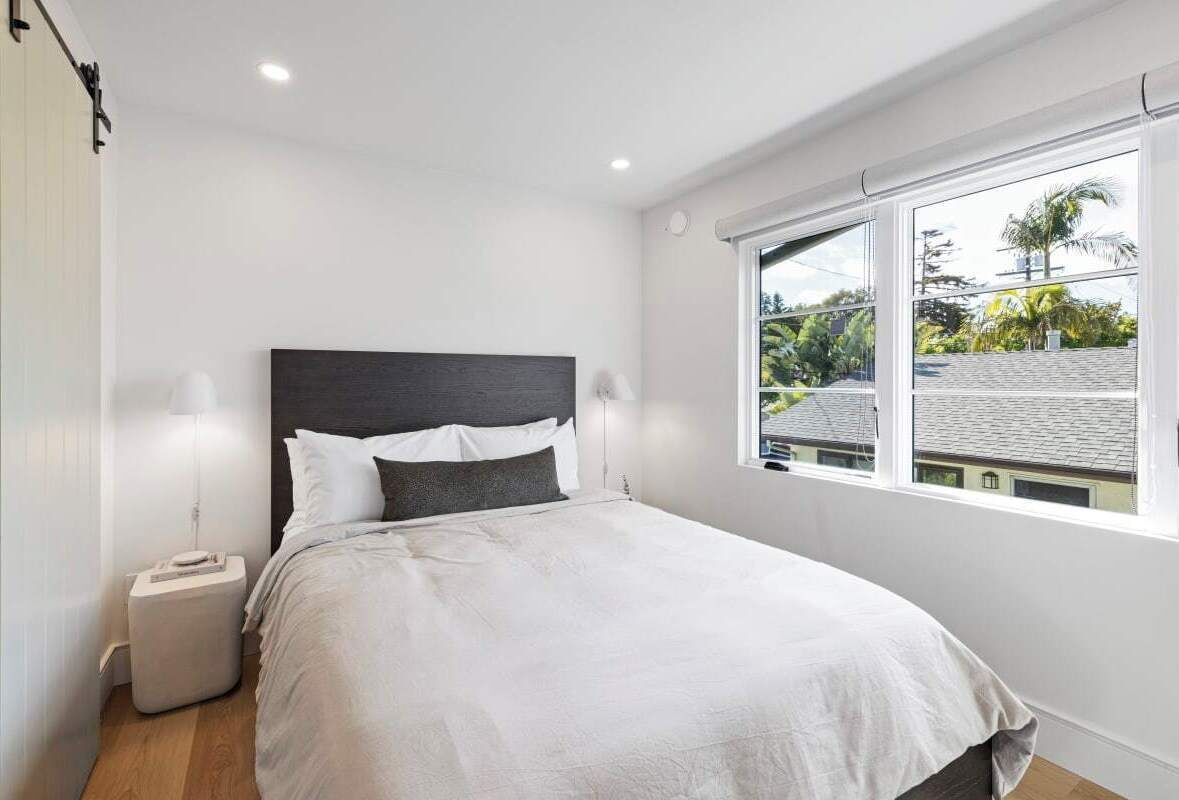 A bedroom with a bed, bedside lamps, a window with a view of buildings and trees, and a white wall.