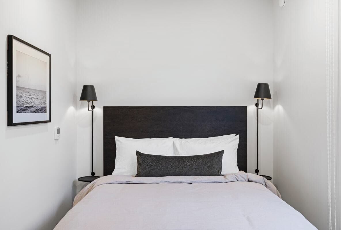 A neatly made bed with white and grey bedding, two black lamps on either side, and a framed picture on the wall.