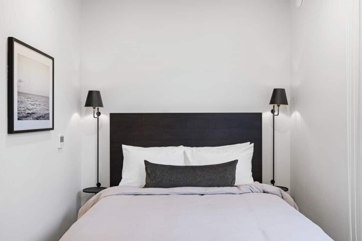 A neatly made bed with white and grey bedding, two black lamps on either side, and a framed picture on the wall.