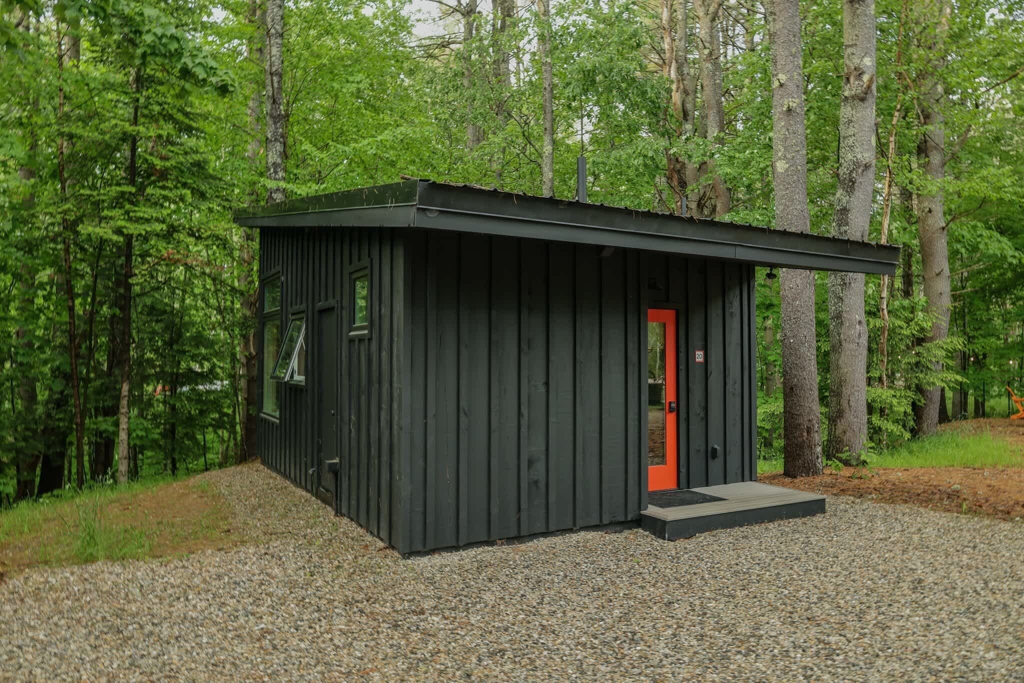 A small, dark wooden cabin with a red door, surrounded by a gravel path and dense forest.