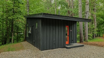 A small, dark wooden cabin with a red door, surrounded by a gravel path and dense forest.