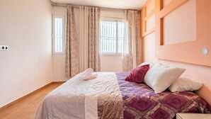 Ein ordentlich bezogenes Bett mit weißer und violetter Tagesdecke, einem weißen Kissen und einem roten Kissen. Ein Fenster mit Vorhängen, ein Holzboden und ein an der Wand befestigter Lichtschalter sind zu sehen.