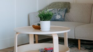 Une table ronde en bois, sur laquelle sont posés un pot blanc et une plante, est placée devant un canapé beige agrémenté d’un plaid vert et d’un coussin bleu.