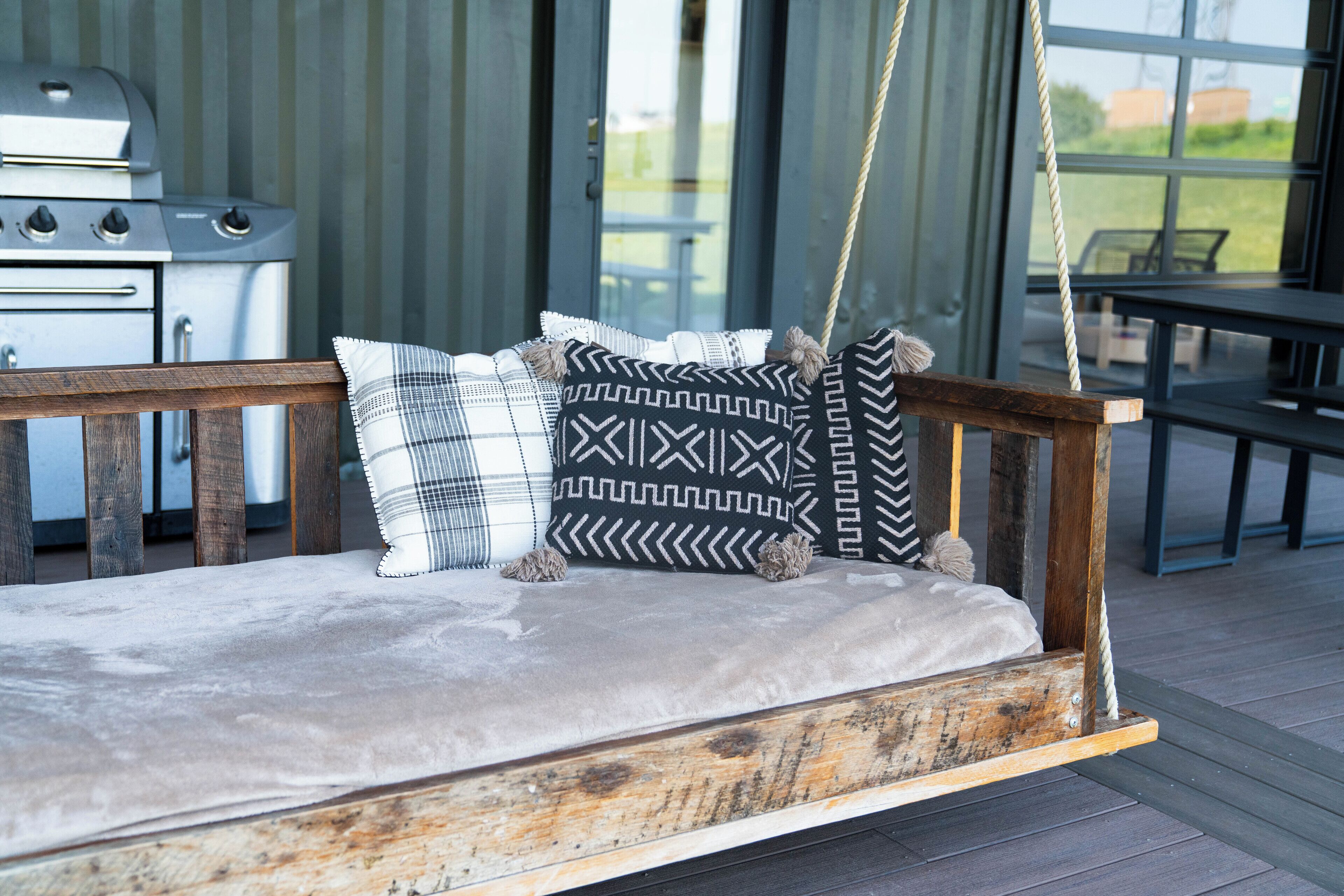 A wooden swing with a cushion and patterned pillows, set on a deck with a grill and dining area in the background.