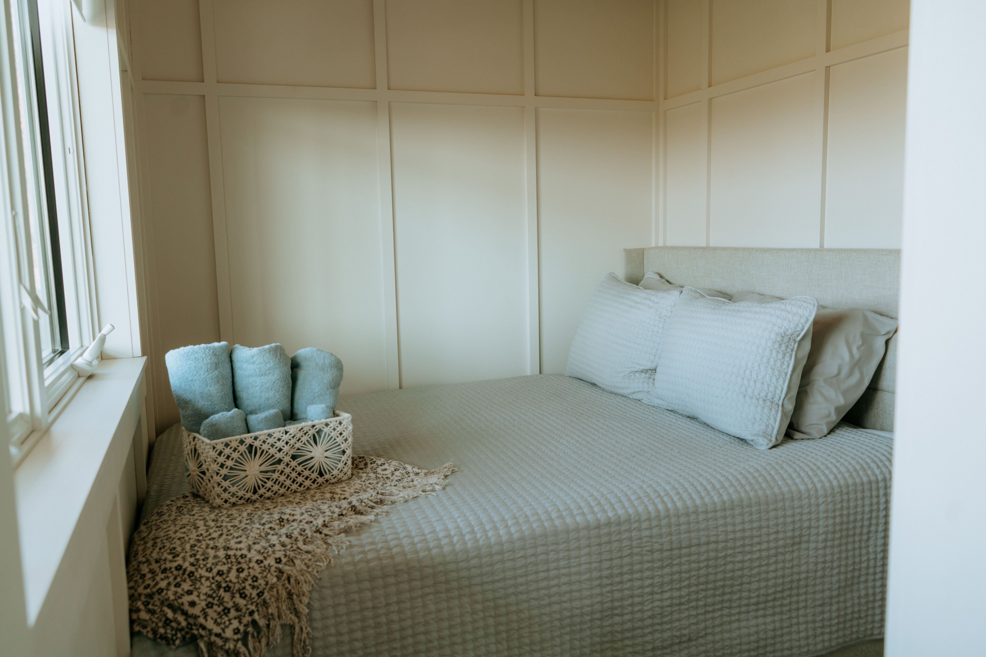 A neatly made bed with a light-colored quilt, a bedside basket with folded towels, and a window with blinds.