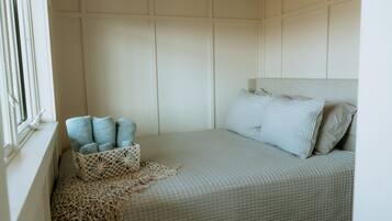 A neatly made bed with a light-colored quilt, a bedside basket with folded towels, and a window with blinds.