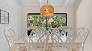 Un comedor con una mesa y sillas blancas, una lámpara colgante de fibras naturales y una ventana con vistas a palmeras.