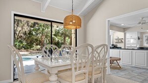 Un comedor con mesa de vidrio y sillas blancas, una lámpara colgante de mimbre y vista a una piscina y follaje.