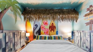 A tropical-themed bedroom with a bed, pillows, and a wall decorated with surfboards and a beach sign.