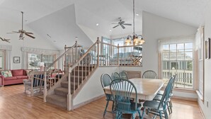 A spacious living area with a wooden staircase, a dining table with chairs, and a large window with a view. - 1285 - Sands to Sea - Pet-Friendly Corolla Retreat Near the Beach (Corolla)