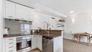 A modern kitchen with white cabinetry, stainless steel appliances, a granite countertop, and a dining area with a wooden table and chairs. - Bright 1 Bedroom Beauty with Balcony Zodiak Stays (Auckland)