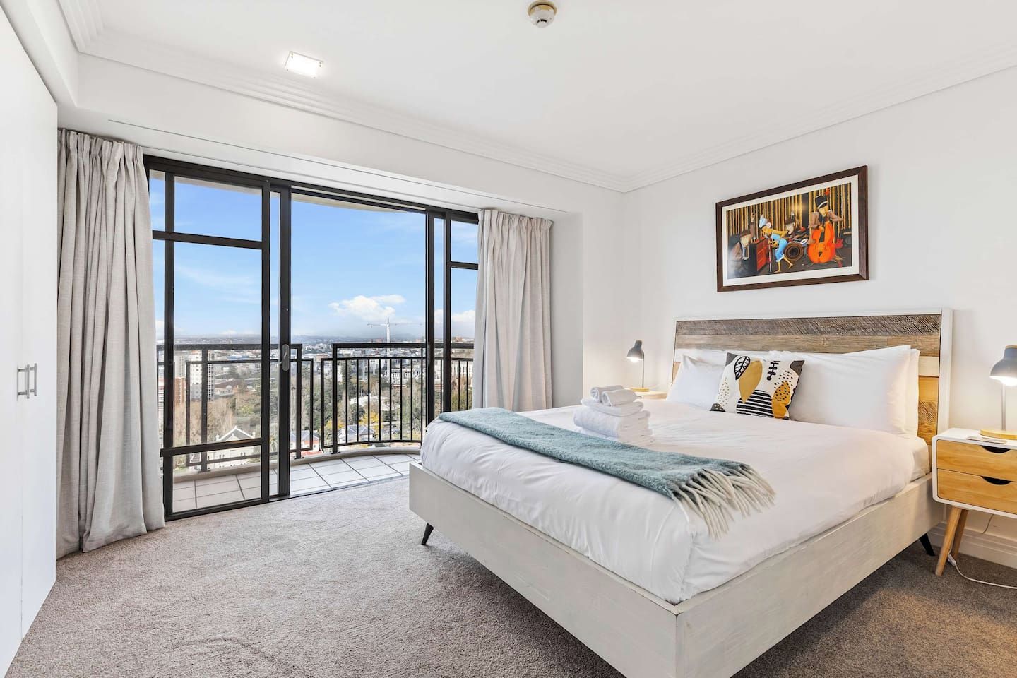 A hotel room with a large bed, a nightstand, a painting, and a view of the city through a balcony.