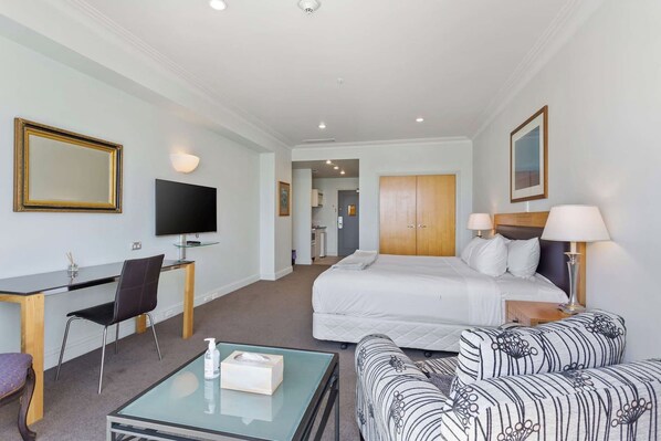 A hotel room with a bed, a desk with a chair, a television, a sofa, and a bedside table with a lamp.