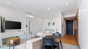 A modern living room with a flat-screen TV, a sofa, a dining table with chairs, and a kitchen area in the background. - Sleek Modern Studio with Balcony by Zodiak Stays (Auckland)