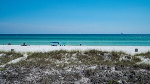 A beach with white sand, clear blue water, and sparse vegetation.