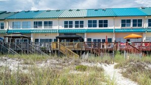 A beachfront hotel with multiple-story buildings, a sandy path leading to the entrance, and a fenced area with a wooden deck.