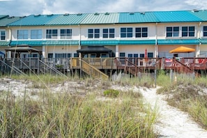 Un hotel frente a la playa con edificios de varios pisos, un camino de arena que conduce a la entrada y un área cercada con una terraza de madera.