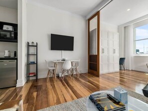 Un salon moderne doté d’un téléviseur à écran plat, d’un coin repas avec une table et des chaises blanches, d’une cuisine équipée d’un four à micro-ondes et d’une machine à café, et d’une grande fenêtre laissant entrer la lumière naturelle.