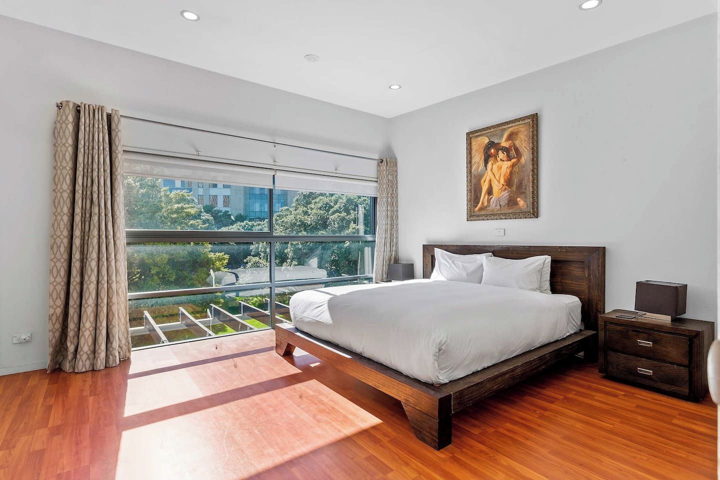 Une chambre moderne avec un grand lit, une table de chevet, un tableau et une vue sur la verdure par une grande fenêtre.