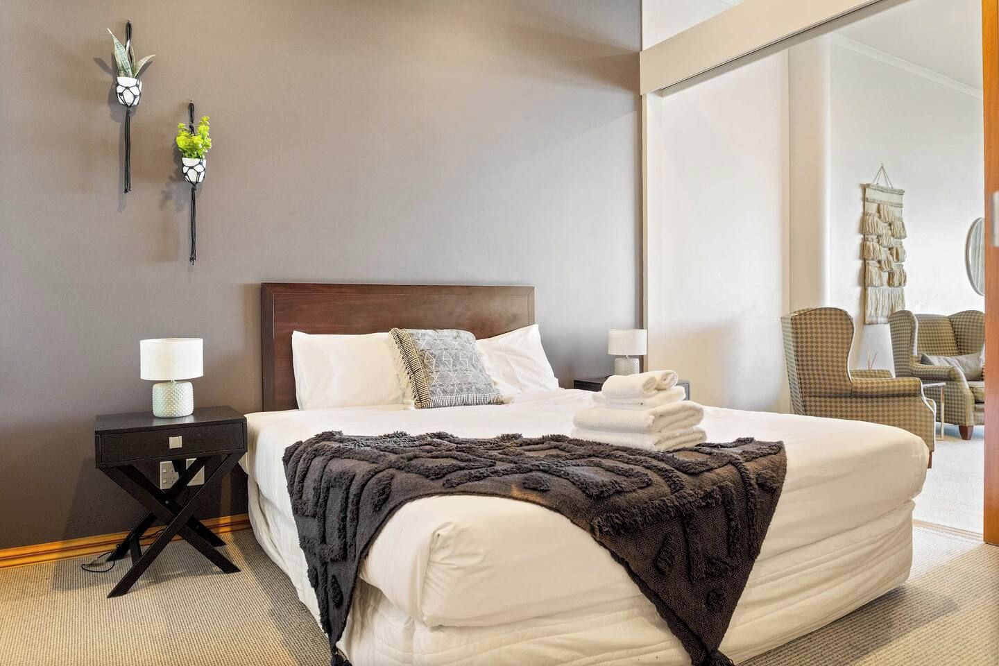 A hotel room with a large bed, two bedside tables with lamps, a dark-colored throw blanket, and a wall-mounted macrame piece.