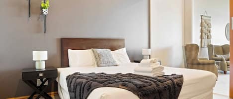 A hotel room with a large bed, two bedside tables with lamps, a dark-colored throw blanket, and a wall-mounted macrame piece.