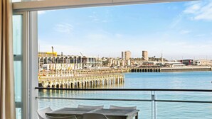A balcony with a table and chairs, overlooking a waterfront with a pier and industrial buildings. - Relaxing Sea Wharf View Apartment by Zodiak Stays (Auckland)