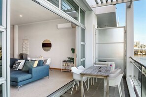 A modern living room with a blue sofa, a dining table with chairs, and a large window offering a view of the city. - Relaxing Sea Wharf View Apartment by Zodiak Stays (Auckland)